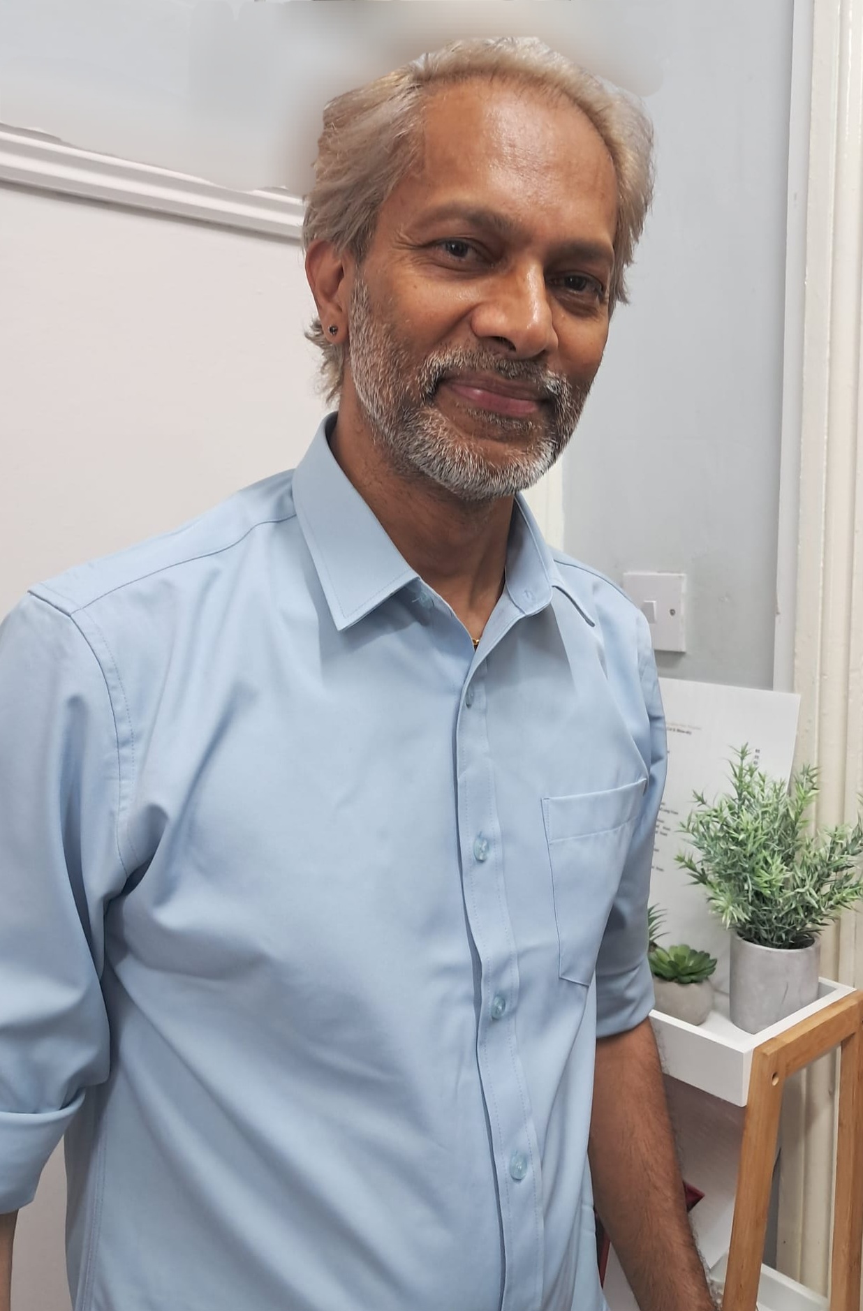 Smiling older man with gray hair and beard wearing a light blue shirt indoors near potted plants.